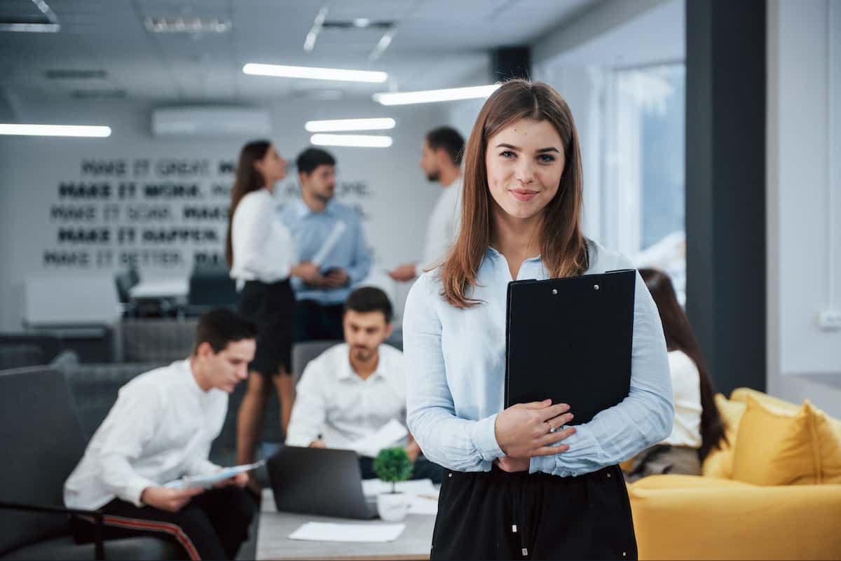 young woman standing with a clipboard in business attire, smiling, in front of blurred office workers in a white room with florescent lights